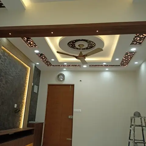 Living room entrance with a multi-tier false ceiling featuring laser-cut jaali insets around a central fan medallion and a dark backlit textured wall panel