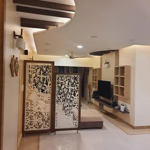 Living room entry framed by a carved floral jaali partition screen under a curved wood-beamed false ceiling, opening to a cubbied TV wall beyond.