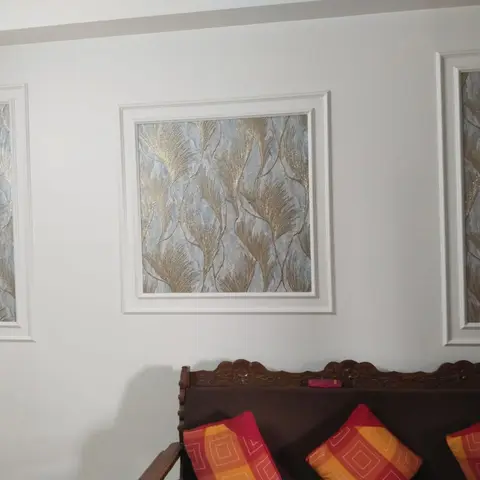 Living room feature wall with white moulded panel frames around gold-and-blue leaf-motif fabric inserts above a carved wood bench with bold cushions.