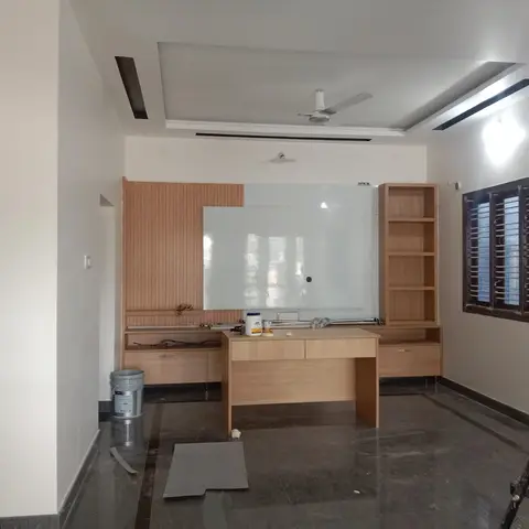 Living room mid-fit-out with a light-wood fluted TV unit and open shelving flanking a reflective center panel, ornately carved wooden main door to the side