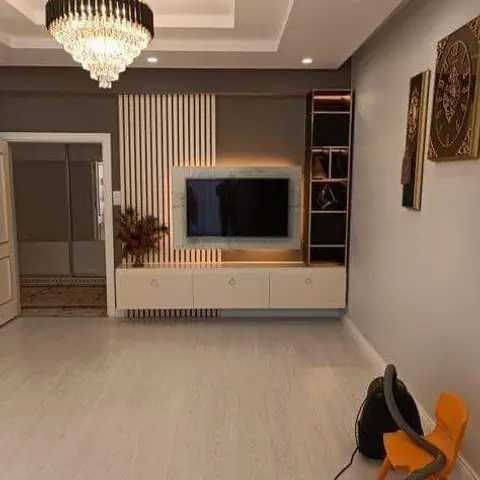 Living room mid-fit-out with a fluted-panel TV wall and crystal chandelier, floor still bare apart from a vacuum cleaner and a child's chair.