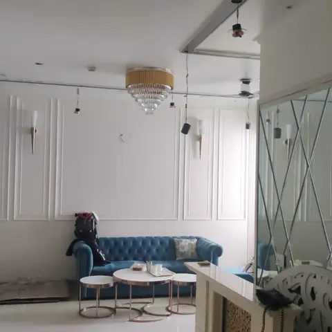 Living room mid-fit-out with a blue tufted chesterfield sofa, fluted wall-panel moulding, a gold drum chandelier and a diamond-pattern mirrored feature wall.