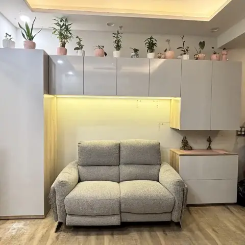 Living room nook with a grey fabric reclining loveseat beneath overhead grey gloss storage cabinets lit with warm cove lighting