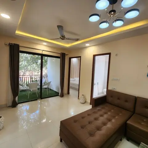 Living room of a flat with a brown buttoned-leather L-sofa under a cove-lit recess holding an eight-globe chandelier and a ceiling fan