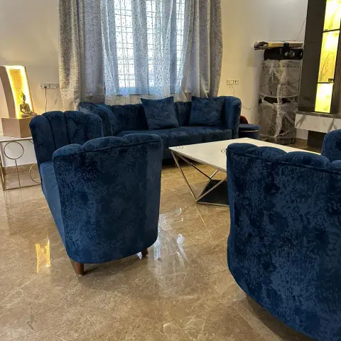 Living room part-way through handover, with a blue crushed-velvet sofa and two tub chairs round a marble-top table on a chrome X-frame