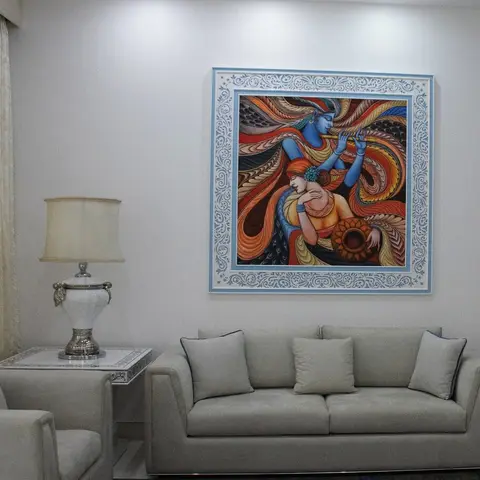 Living room seating area with a large framed Radha-Krishna painting above a cream sofa, an ornate silver table lamp on a mirrored side table