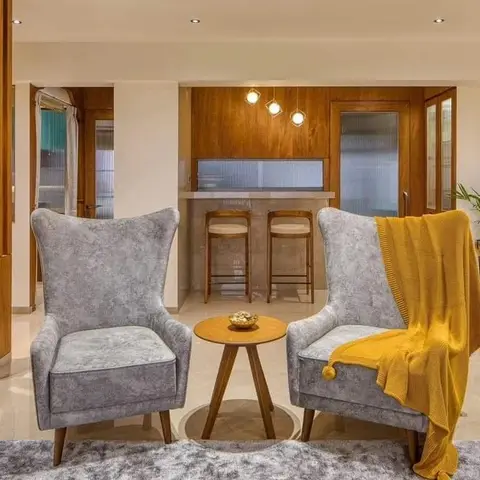 Living room seating nook with two grey wingback armchairs and a mustard throw, framed by a wood-panelled home bar with pendant lighting beyond.
