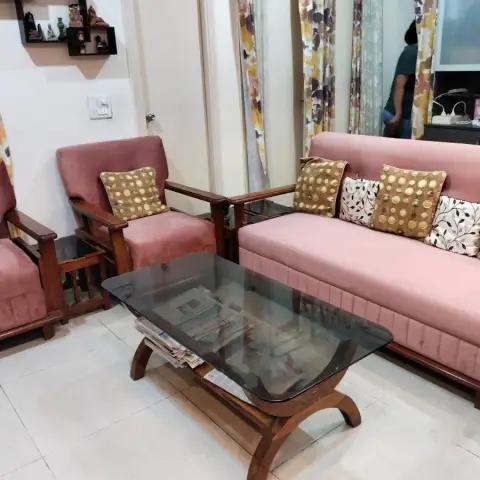 Living room seating set with wood-framed pink upholstered sofa and armchairs around a glass-top coffee table stacked with newspapers.