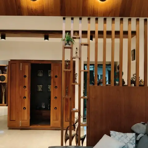 Living room seen past a grey sofa, where a solid-teak vertical slat screen with staggered shelves of brass figurines stands beside an open teak pooja cabinet