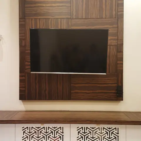 Living room TV feature wall made of patchwork wood-veneer panels with a mounted TV, above a floating console with geometric jaali-pattern cutwork inserts.