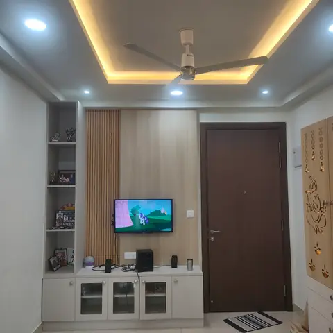 Living room TV feature wall with vertical wood fluted panelling and a cove-lit ceiling, beside a laser-cut jaali Ganesh motif panel near the main door.