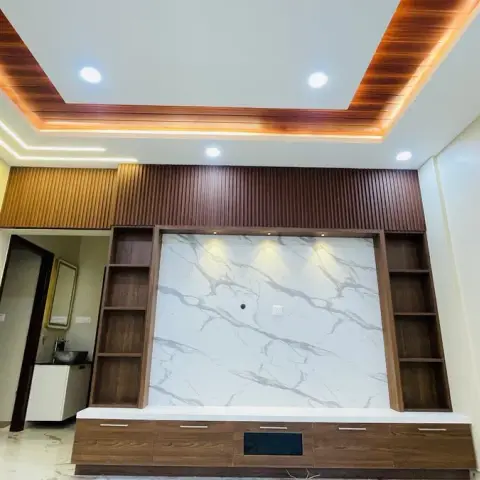 Living room TV feature wall with a book-matched white marble panel framed by dark wood fluted panelling and open shelving, cove-lit wood ceiling drop above