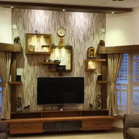 Living room TV unit flanked by curtained bay windows, with a textured wallpaper backdrop and illuminated wood display niches showcasing curios and a wall clock.