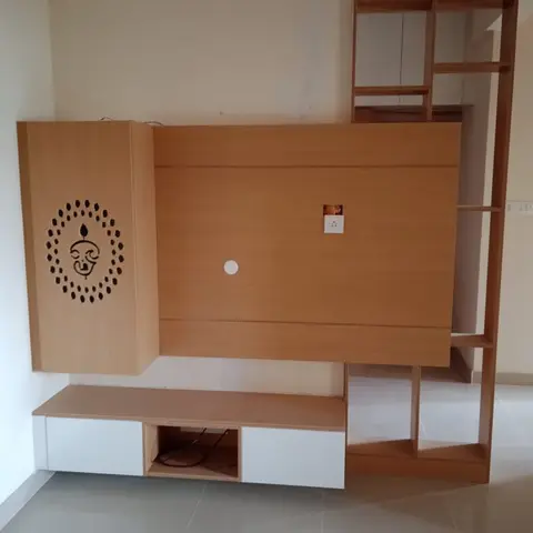 Living room TV unit in a light-oak laminate finish with a Ganesha-motif CNC jaali side panel and an open ladder display rack, still awaiting wall finishing.