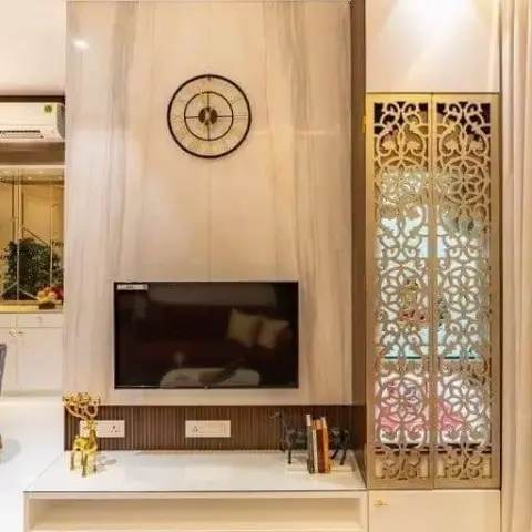 Living room TV wall in cream marble with a round wall clock, flanked by a tall cabinet with a gold jaali-pattern glass door, over a white floating TV console