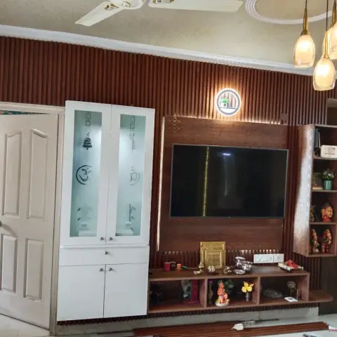 Living room TV wall in fluted wood panelling with a built-in frosted-glass mandir cabinet beside the console displaying idols.