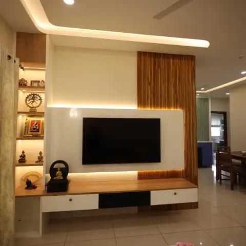 Living room TV wall lit by a backlit white panel, with a teak fluted screen wrapping its right edge and a column of LED-lit niches on the left holding a brass