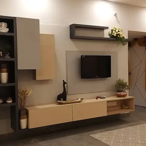 Living room TV wall on a housewarming day: beige-and-charcoal laminate console with a floating shelf and open display boxes holding brass curios and Ganesha