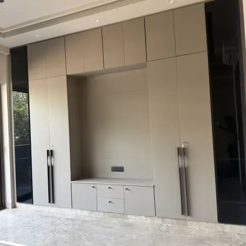 Living room TV wall unit with beige lacquer joinery, a recessed TV niche, tall bar-pull handles and marble flooring beside a glass balcony door