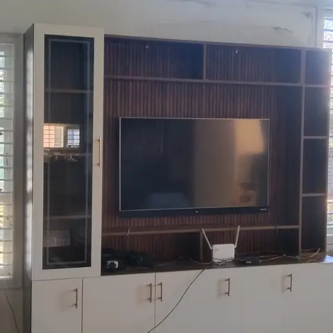 Living room TV wall unit with a wood-slat backdrop and glass-front display cabinet, white base cabinets, facing a pair of wicker chairs.