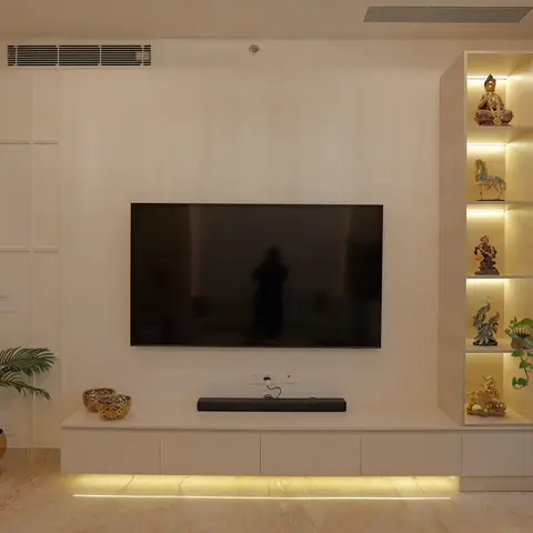 Living room TV wall with a floating ivory console lit by a warm floor-level strip light and a backlit display niche of brass and stone curios beside the