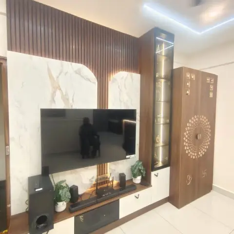 Living room TV wall with a marble slab backdrop, wood fluted panelling, a lit glass display shelf, and an adjoining wood pooja cabinet with a brass jaali motif.