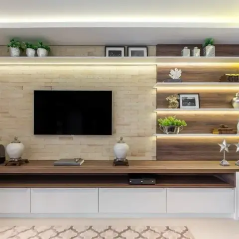 Living room TV wall with beige stone cladding, cove lighting, and a backlit wooden display shelf beside a floating wood-and-white TV unit.