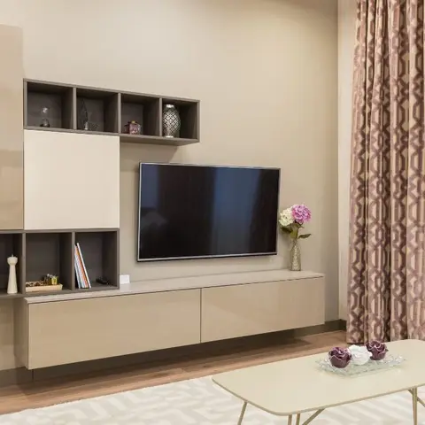 Living room TV wall with floating taupe gloss joinery and open display cubbies styled with books and vases, beside a patterned drape and an oval coffee table.
