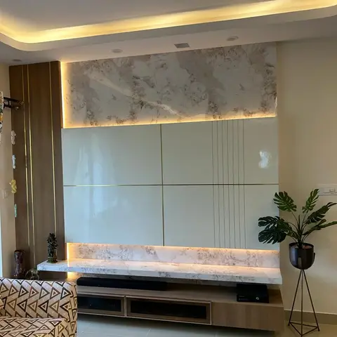 Living room TV wall with marble-clad top and bottom bands framing a glossy gold-lined panel, beside a fluted wood pillar and a monstera plant on a metal stand.