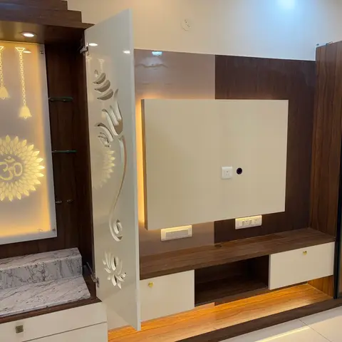 Living room wall unit combining a backlit pooja nook with an Om motif and hanging bells beside a TV unit wall with a glass-fronted display cabinet.