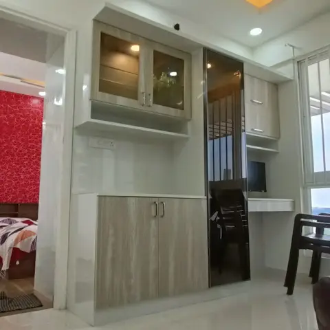 Living room wall unit with a light wood-grain glass-front cabinet and dark mirrored door, opening onto an adjoining bedroom with red patterned wallpaper.