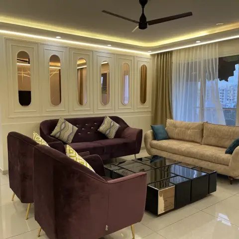 Living room where a cream moulded panel wall is punched with a row of capsule-shaped bronze mirror insets behind a plum velvet sofa and tub chairs