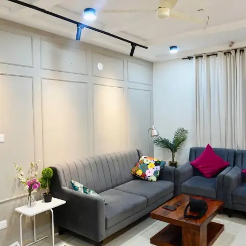 Living room where a grid of slim applied mouldings covers the long greige wall, lit by a black ceiling track of spots