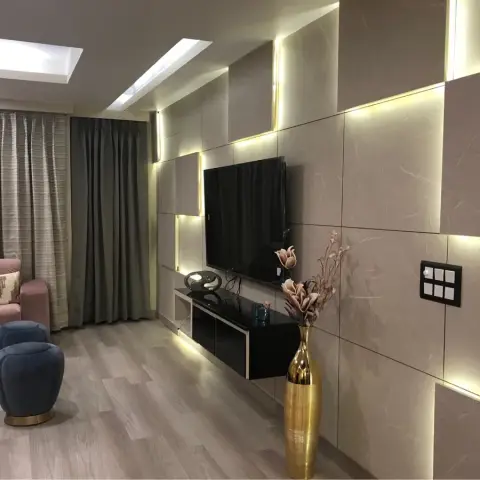 Living room where a staggered beige stone-effect panel wall is lit by strips hidden in its setbacks, with a black floating TV console