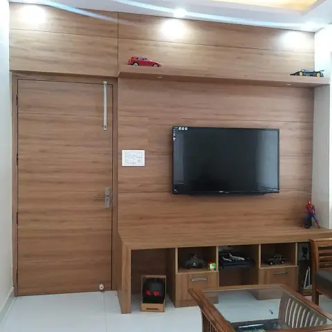 Living room where a teak-laminate wall runs unbroken from the flush door across the TV panel