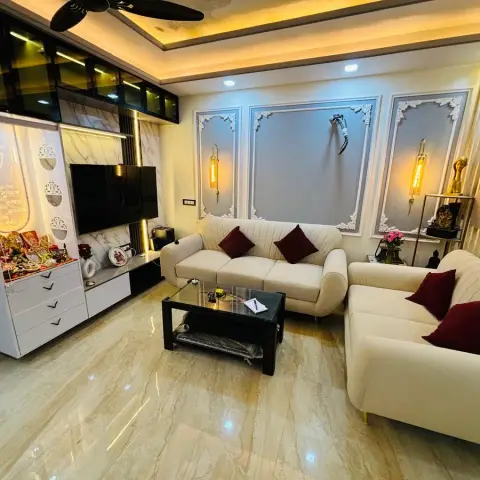Living room whose end wall combines a backlit white jaali pooja unit with an Om cut-out and deity idols, a TV on marble-look cladding and glass-fronted lofts
