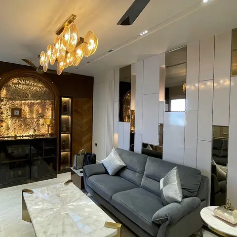 Living room with a backlit amber-stone bar niche, chevron wood panelling, and a sculptural glass-and-brass chandelier over grey velvet sofas.