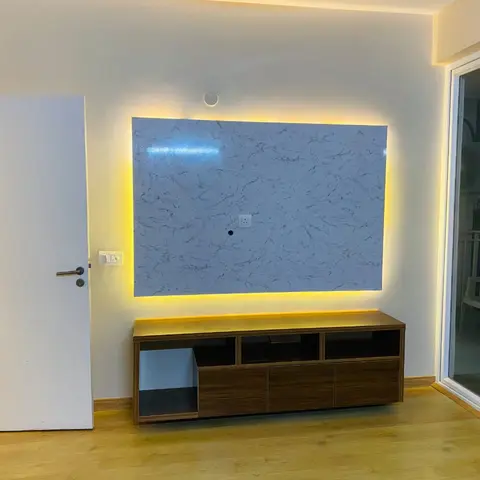 Living room with a backlit grey-veined marble TV feature wall in a warm LED glow, a walnut TV console below, and a sliding door opening onto the balcony.