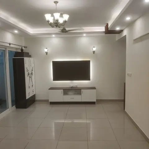 Living room with a backlit TV feature wall, a chandelier under a stepped false ceiling, and a tall pooja cabinet by the balcony door