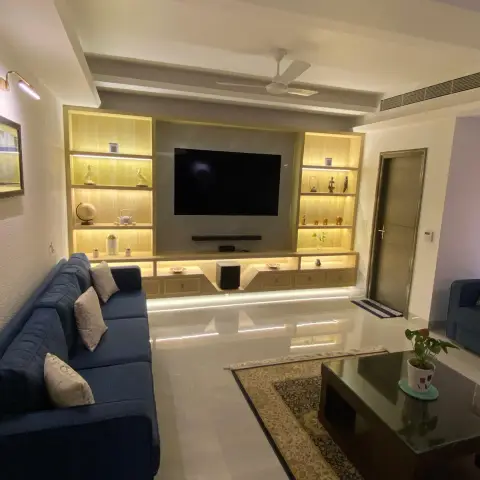 Living room with a backlit wood-toned TV shelving wall styled with curios, a navy sofa, and a floral rug under a glass coffee table.
