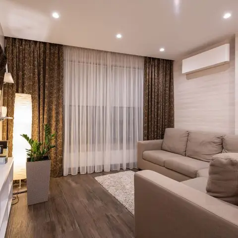 Living room with a beige sectional sofa, layered brown patterned drapes over sheers, wood-look wall cladding and a tall potted plant beside a floor lamp.