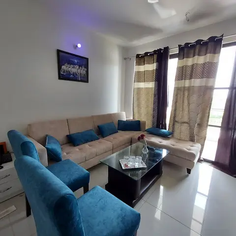 Living room with a beige sectional sofa and teal accent armchair around a glass-top coffee table, patterned drapes framing the balcony door.