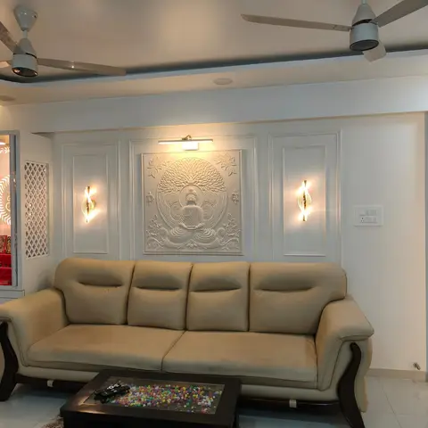 Living room with a beige tufted sofa beneath a carved Buddha relief wall panel flanked by wall sconces, with a jaali-screened pooja unit to the side.