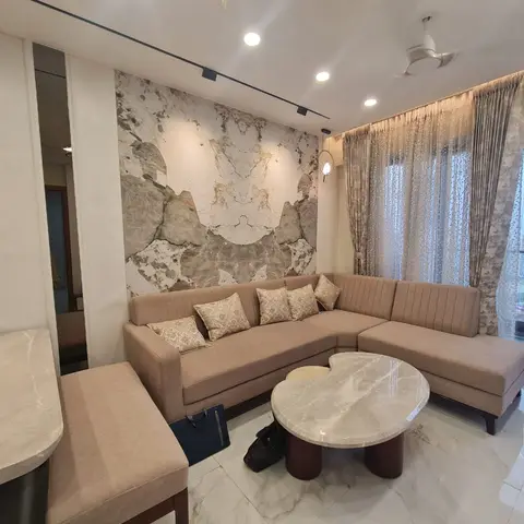 Living room with a beige tufted L-shaped sofa and gold cushions against a marble-effect feature wallpaper, a round marble coffee table, sheer floral curtains