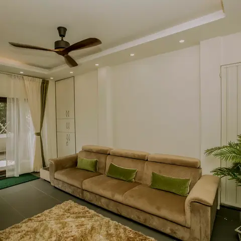 Living room with a beige velvet sofa styled with olive-green cushions, a cove-lit false ceiling and sheer curtains opening to a balcony.