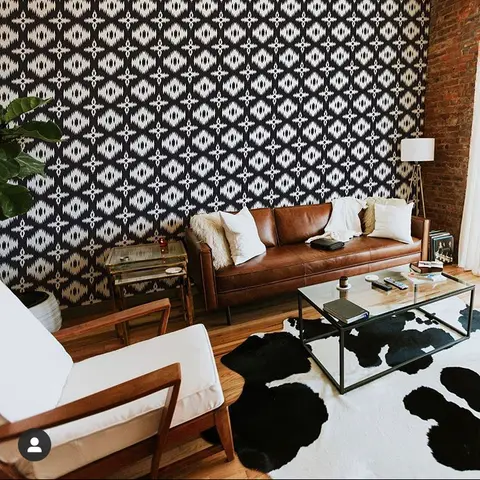 Living room with a bold black-and-white geometric wallpaper accent wall, a tan leather sofa, and a cowhide rug on wood flooring.