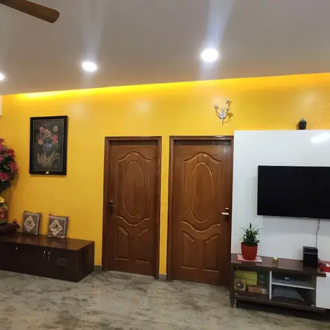 Living room with a bold mustard-yellow cove-lit accent wall, twin varnished wood-panel doors, and a white-and-wood TV unit with a glass display cabinet