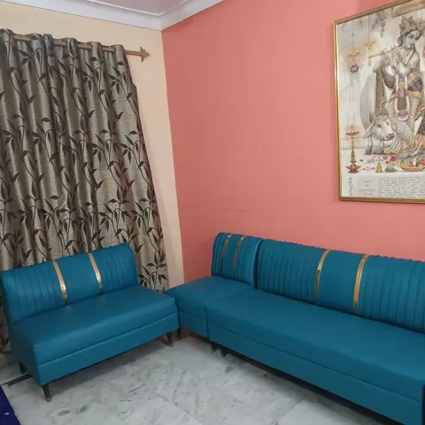Living room with a bold teal channel-tufted L-shaped sofa trimmed in gold, a coral accent wall, a framed religious portrait, and leaf-print curtains.