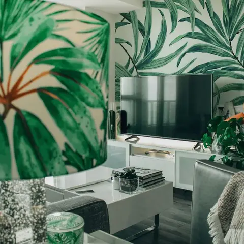 Living room with a bold tropical palm-leaf wallpaper behind a wall-mounted TV, white gloss console, and a grey sofa styled with plants and a flamingo ornament.