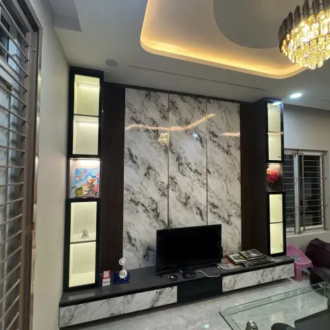 Living room with a book-matched marble TV feature wall flanked by lit glass-front display towers, a curved cove-lit false ceiling with a crystal chandelier